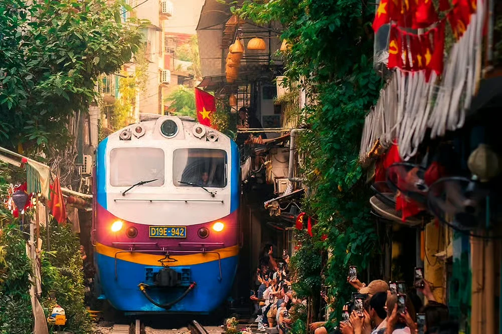 A train passing through a narrow Hanoi street lined with buildings, greenery and Vietnamese flags.